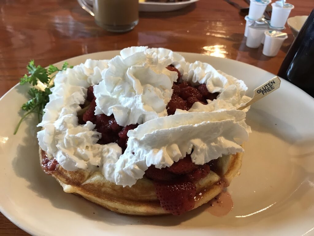 A huge waffle with whipped cream and strawberries. It has a toothpick sticking out of it that says gluten free.