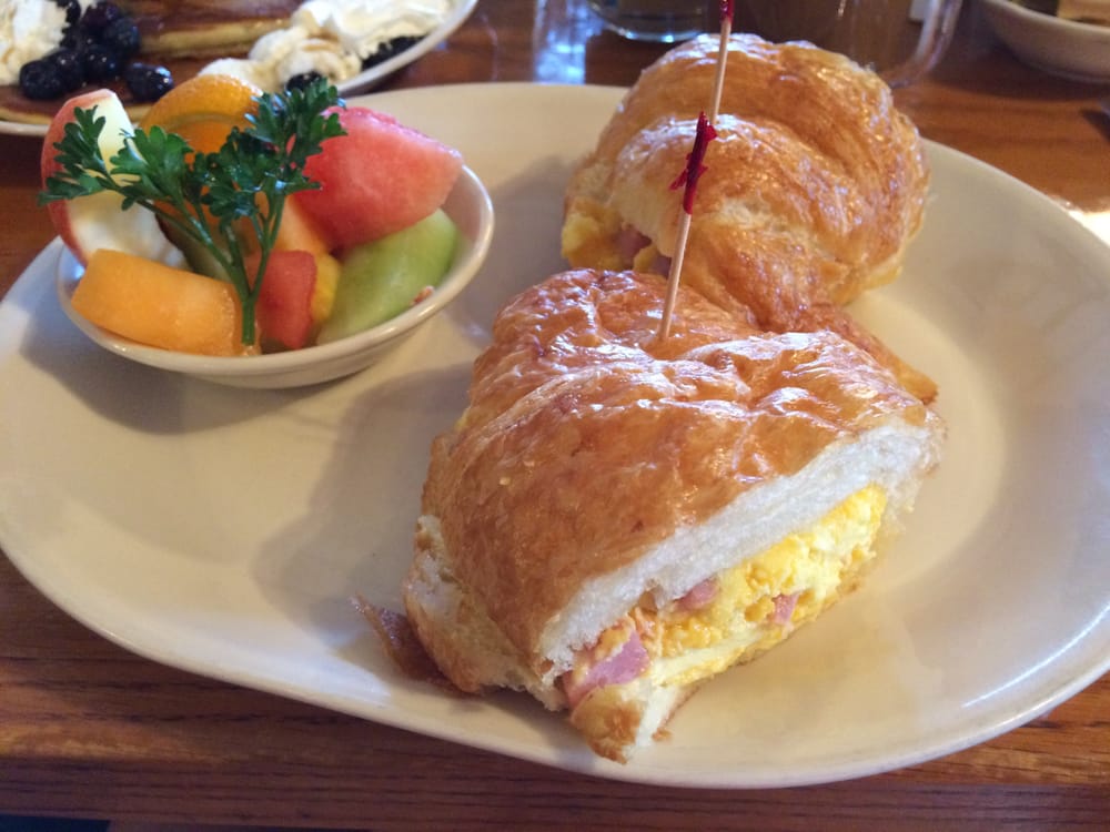 An egg and ham croissant sandwich with a bowl of fresh fruit on a white plate. Red toothpicks are holding the sandwich together.