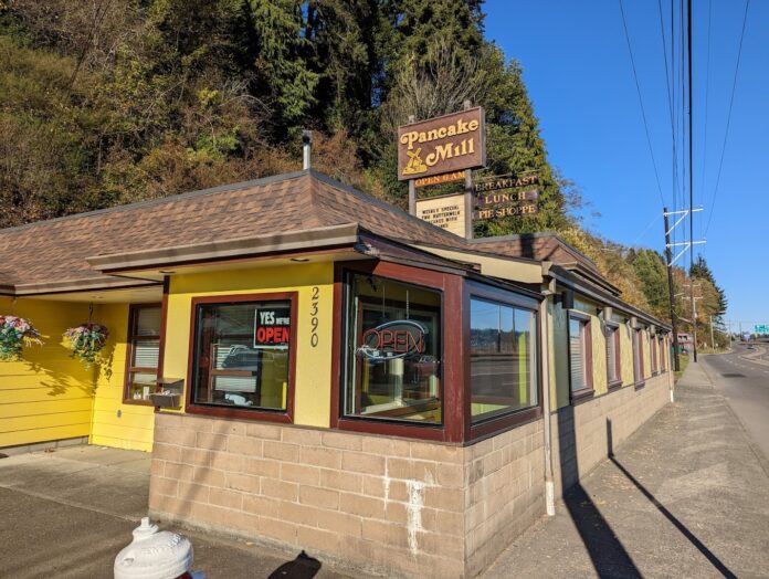 The outside of The Pancake Mill in North Bend, Oregon. The building is yellow, with brown trim and a brown roof. The sign is brown with yellow lettering.