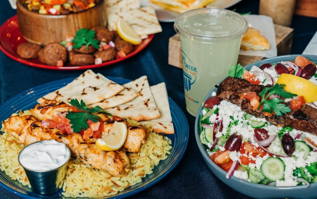 Three plates of colorful Greek food and a yellow drink in a plastic cup.