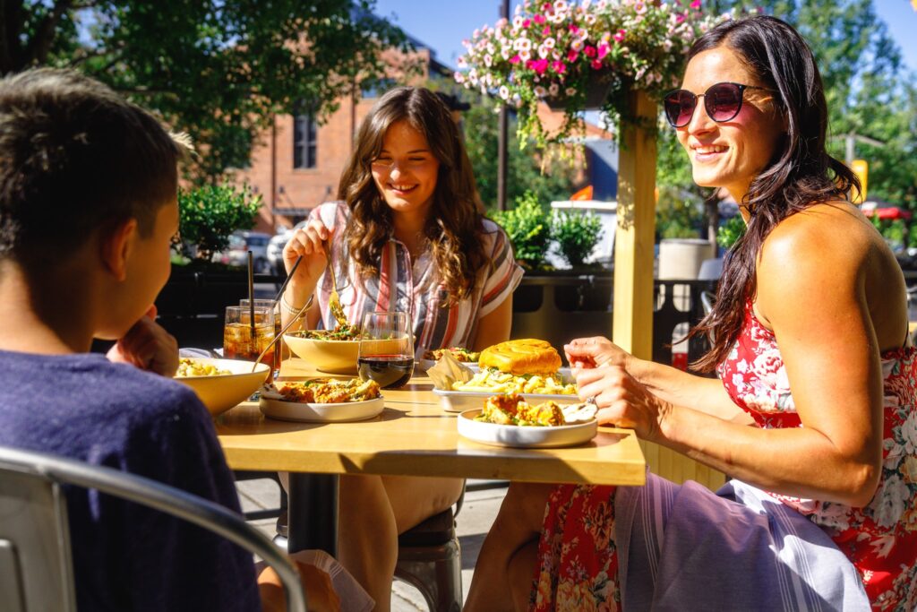 Three people smile and eat at an outdoor table.