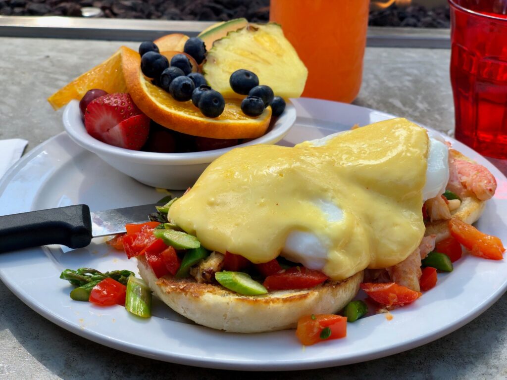 A colorful looking eggs benedict with a bowl of fruit including an orange slice, strawberries, blueberries and pineapple.