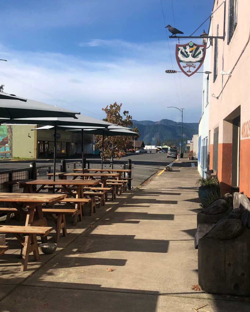 The outside of The 3 Legged Crane in Oakridge, Oregon. There are picnic tables under umbrellas on the sidewalk, and a view of the forested mountains in the distance.