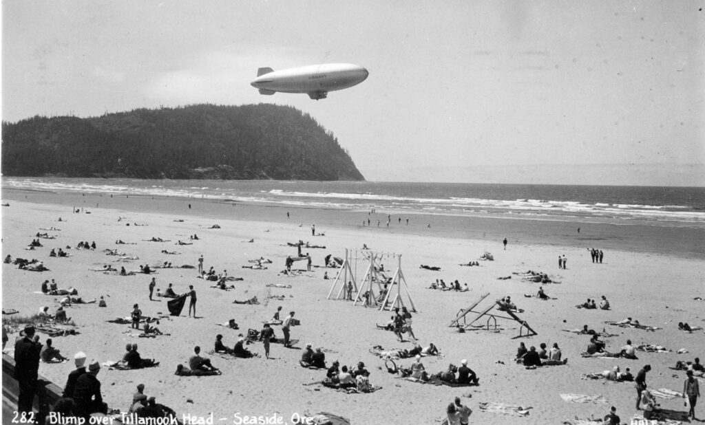 A black and white photo of a K ship flying over the water in Seaside Oregon.  Beachgoers look on. tillamook air museum, planes, WWII, history, family things to do, oregon coast