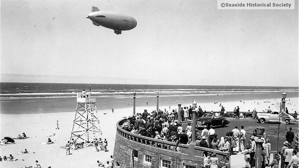 A black and white photo of a K ship flying over the water in Seaside Oregon.  Beachgoers look on. tillamook air museum, planes, WWII, history, family things to do, oregon coast