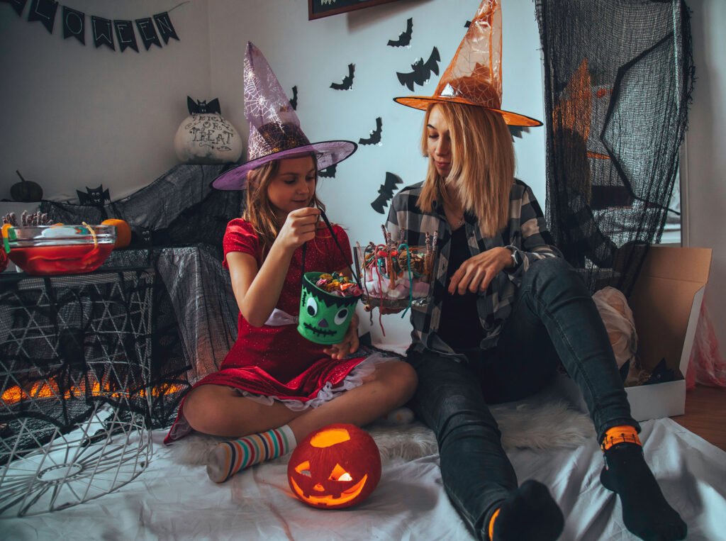 A mother and daughter lookin through Halloween candy, sitting on a bed wearing witch hats.