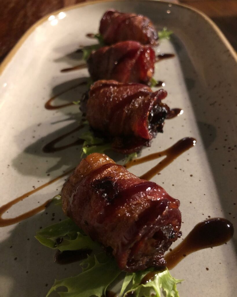 Four bacon wrapped dates lined up on a plate and drizzled with sauce.