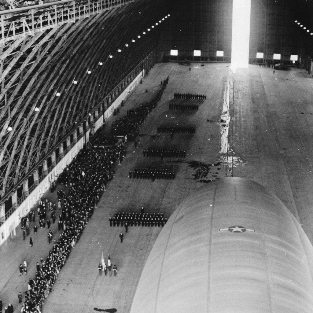 A black and white photo of inside Hangar B during an open house on July 4th, 1945.  People line up and look at a massive blimp inside. tillamook air museum, planes, WWII, history, family things to do, oregon coast