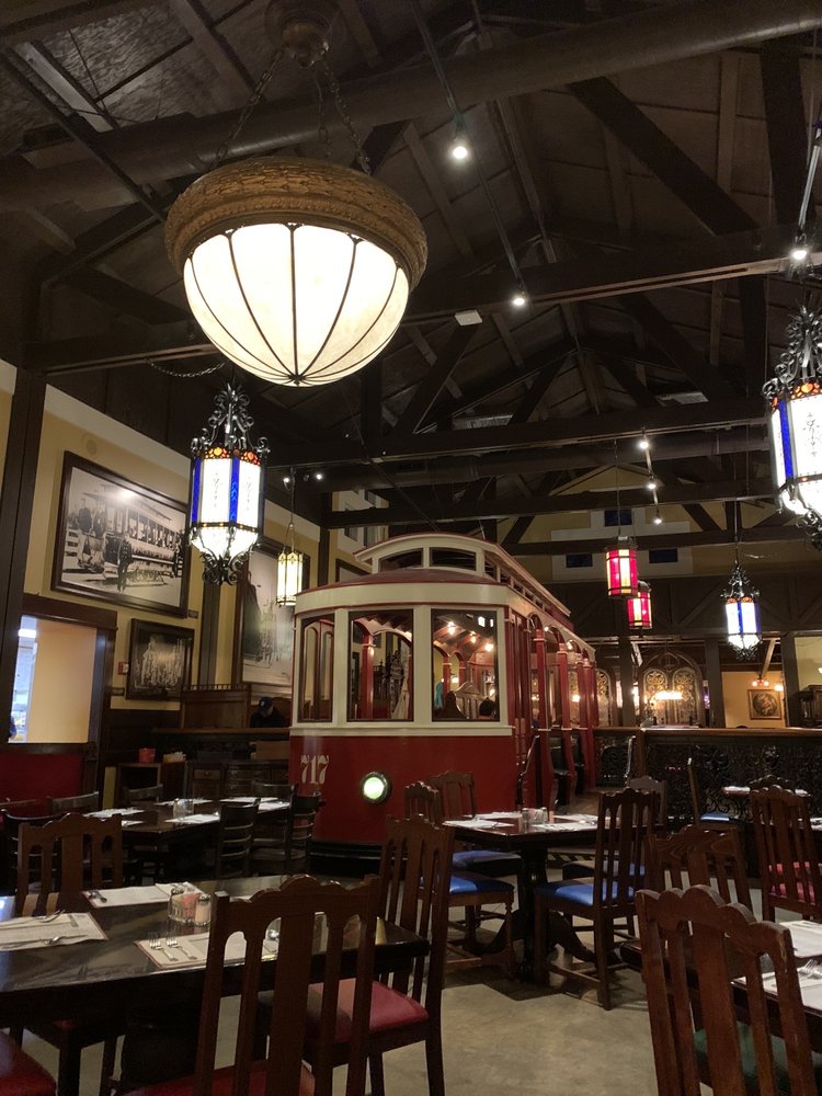 The interior of The Old Spaghetti Factory. There are wooden tables and chairs, an old full size red and white trolley, pretty antique lighting, and wooden beams on the ceiling.