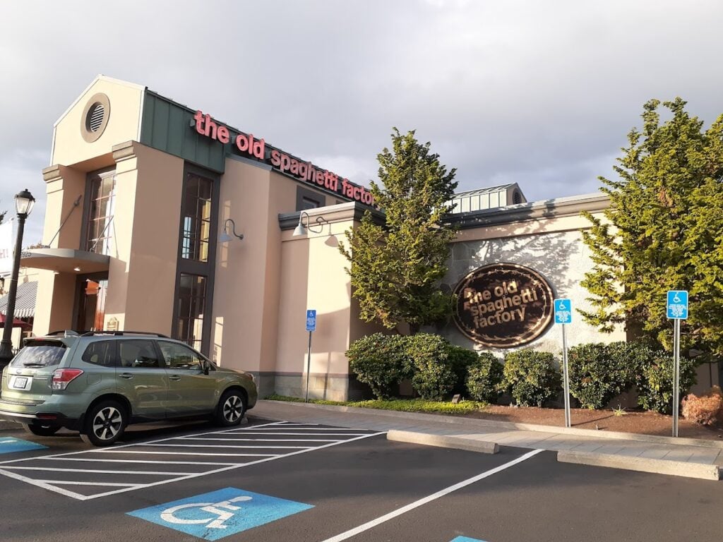 The outside of The Old Spaghetti Factory in Hillsboro, Oregon. It's a tall white building with a green metal roof. there are shrubs and small trees outside.