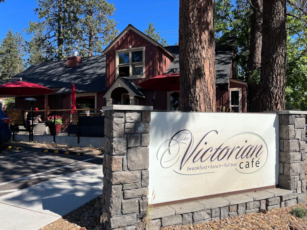 The outside of the Victorian Café in Bend, Oregon. It's a red building with a black roof and a white sign with stone out front. The building is surrounded by tall pine trees.