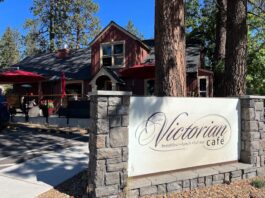 The outside of the Victorian Café in Bend, Oregon. It's a red building with a black roof and a white sign with stone out front. The building is surrounded by tall pine trees.