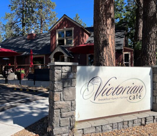 The outside of the Victorian Café in Bend, Oregon. It's a red building with a black roof and a white sign with stone out front. The building is surrounded by tall pine trees.