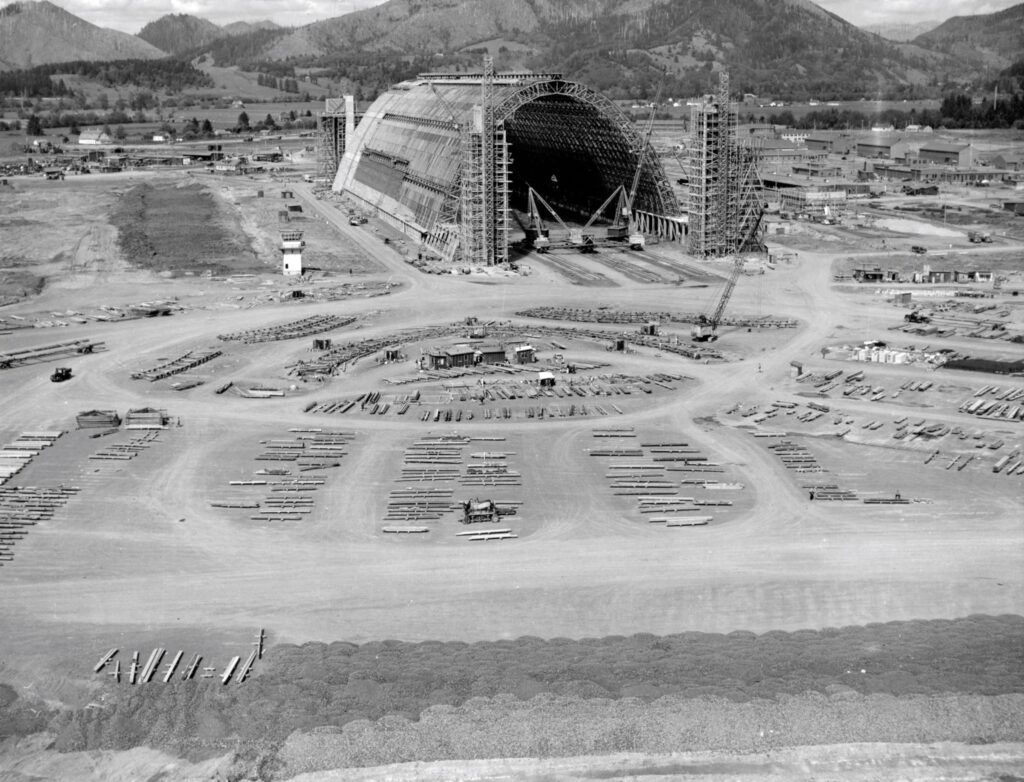 A black and white photo of massive Hangar B being constructed.  There are organized piles of lumber all around the property waiting to be used in construction of the hangar. tillamook air museum, planes, WWII, history, family things to do, oregon coast