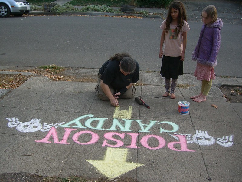 A woman and two children draw the words Poison Candy with an arrow in sidewalk chalk.