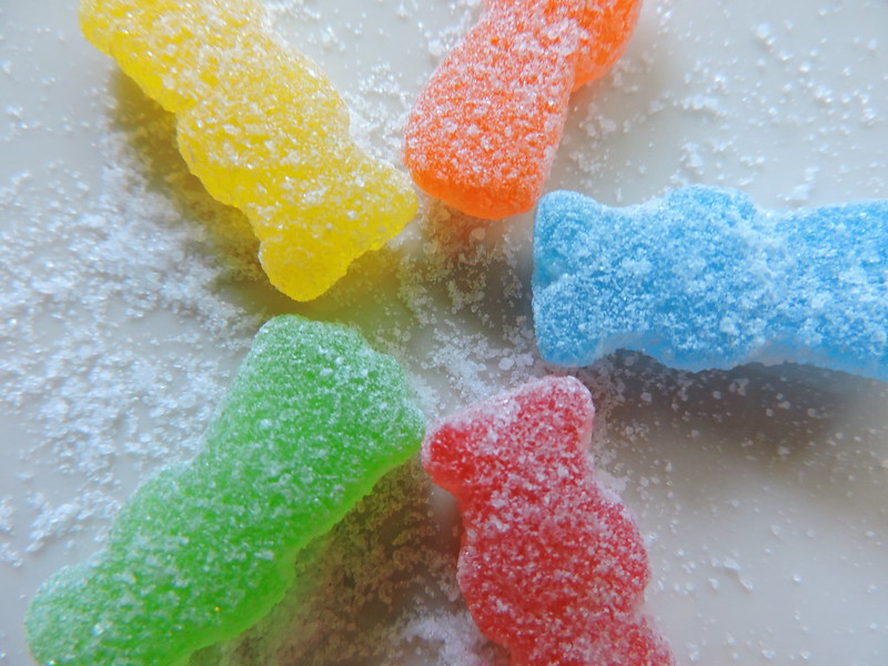Five brightly colored Sour Patch kids candy surrounded by a dusting of sugar on a white surface.