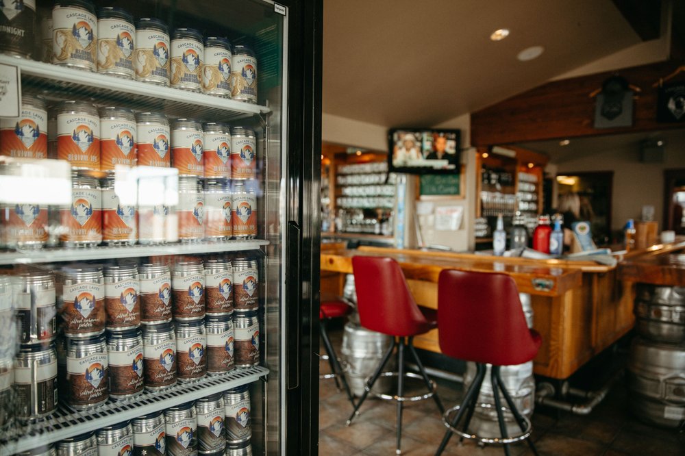 The interior of Cascade Lakes Pub 7th Street Redmond. There's a fridge with cans of beer, warm honey colored wood tables and red chairs.