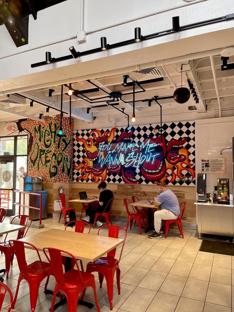 The interior of Dave's Hot Chicken in Eugene, Oregon. Two people sit at wooden tables on red chairs. There is a black and white checkered painting on the wall with graffiti flames.