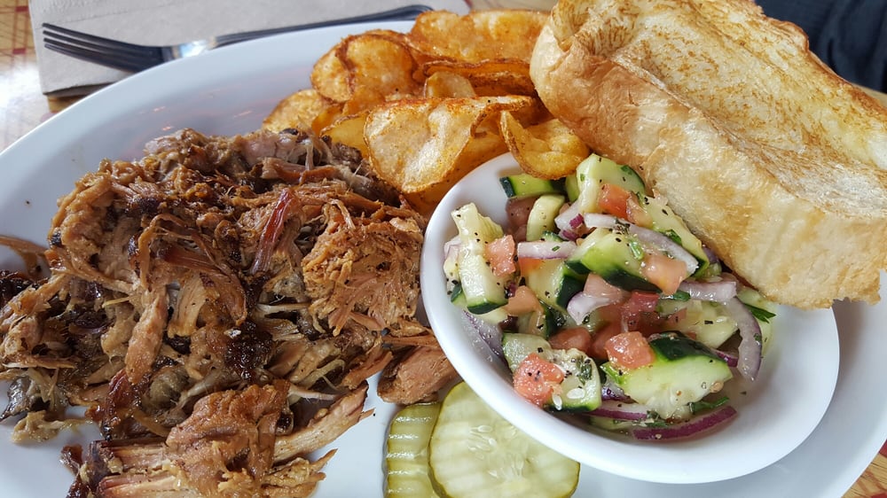 A plate of food with BBQ chips, cucumber salad, pickle slices and shredded meat.