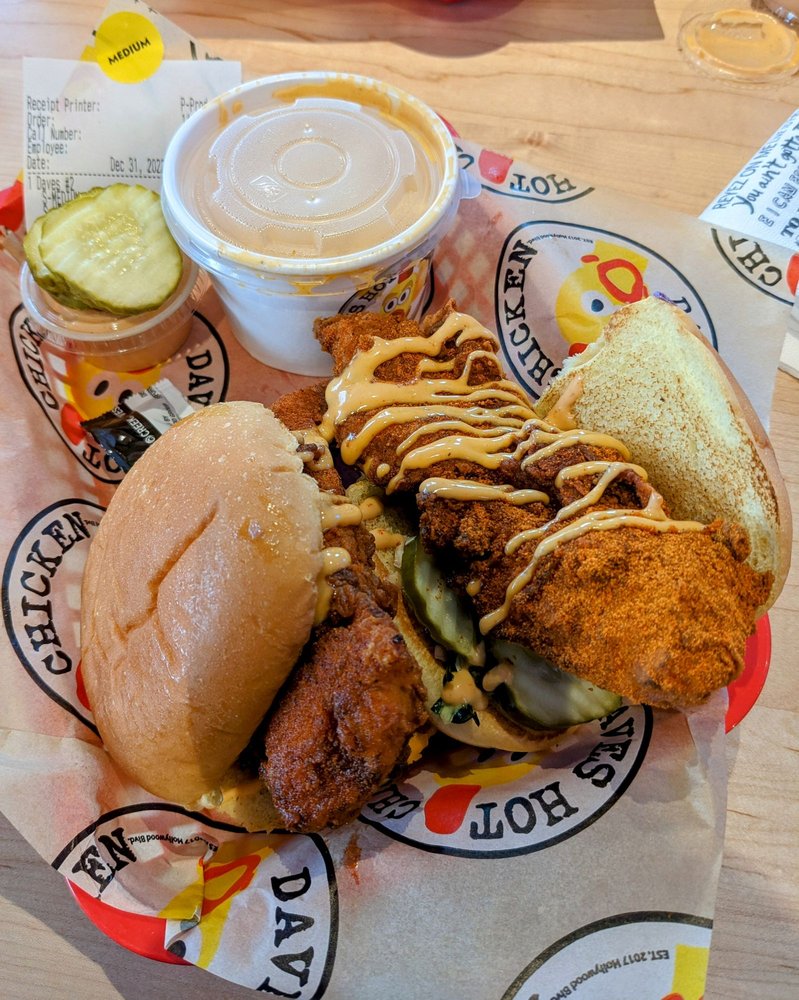 A chicken slider at Dave's Hot Chicken in Eugene, Oregon. There's a side of mac and cheese in a plastic bowl with a lid, cheese drizzled on top of the chicken on the slider, a plastic cup of sauce, and a couple of slices of pickels.