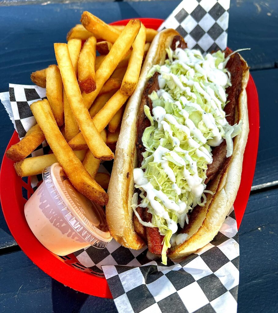 A delicious looking meat sub with lettuce and dressing drizzled over top in a red basket with a black and white checkered paper, fries, and a container of fry sauce.
