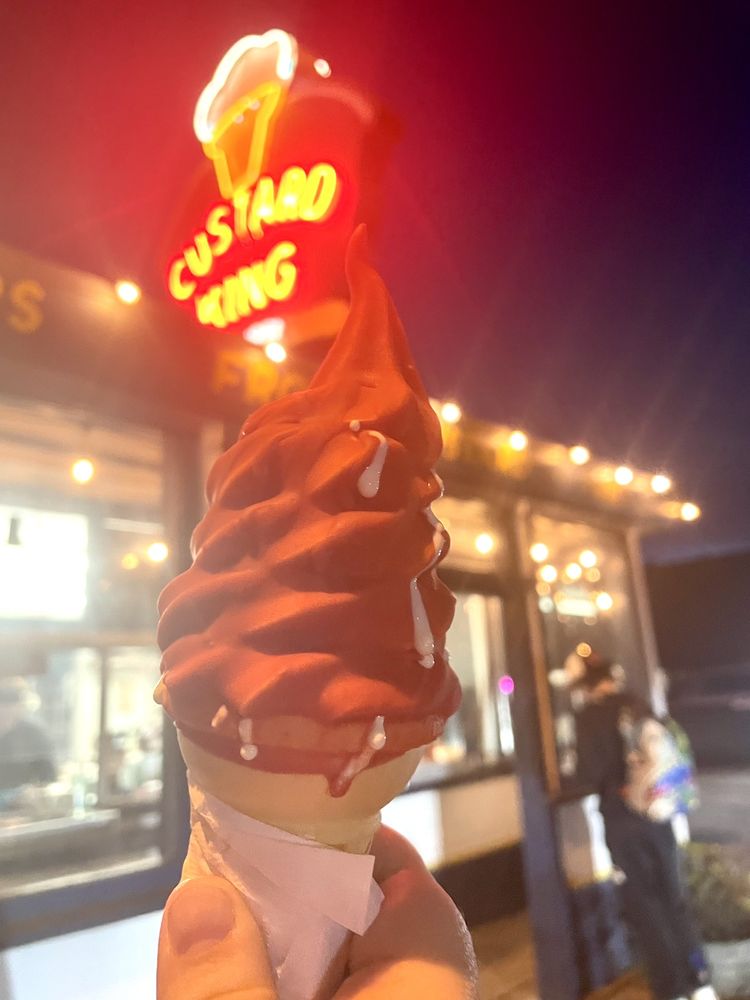 A delicious frozen custard in a cone, held up in front of the neon red Custard King sign at night. The sign is glowing red.