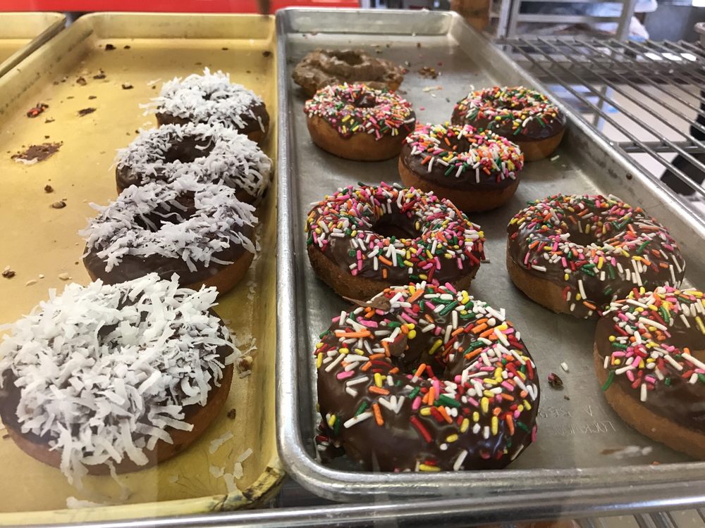 Two kinds of donuts on display in the glass display case. One is chocolate with rainbow sprinkles, the other has shredded coconut on top.