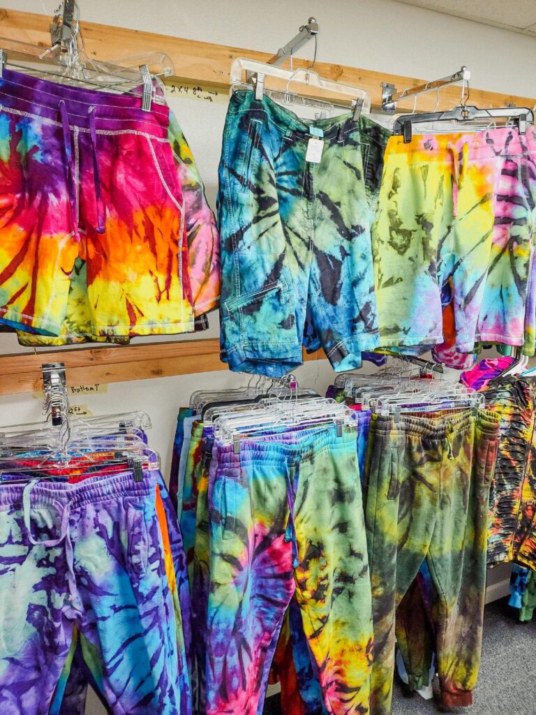 A wide variety of tie die shorts hang on a wall in various colors. There's a striking pair that is purple, white and blue, and another pair that is bright pink, purple, yellow and orange with a splash of blue and green, it looks like a sunset.