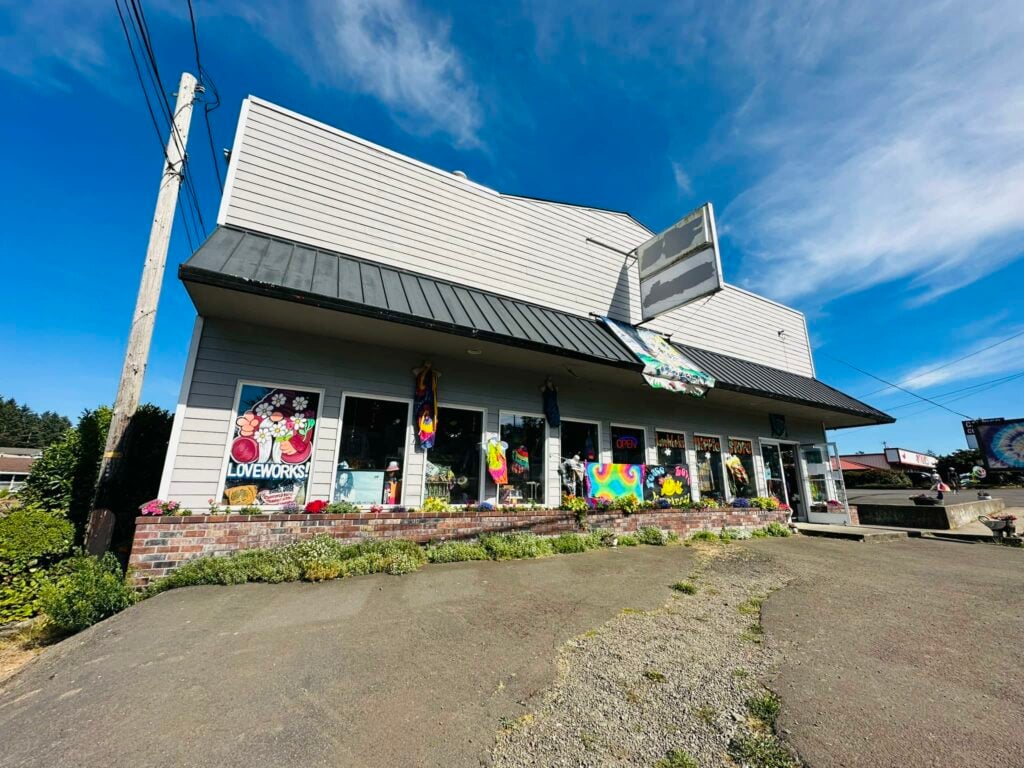 The outside of Love Works Hippie Store in Lincoln City, Oregon. It's a light gray building with a variety of colorful garments and tie dyed fabrics displayed in the windows.