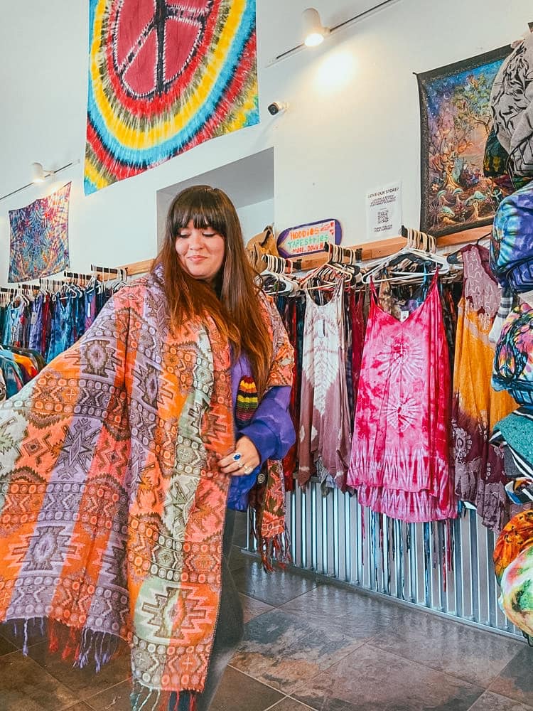 A woman shows off a gorgeous piece of fabric, with a variety of tie dye garments hanging on the wall in the background.
