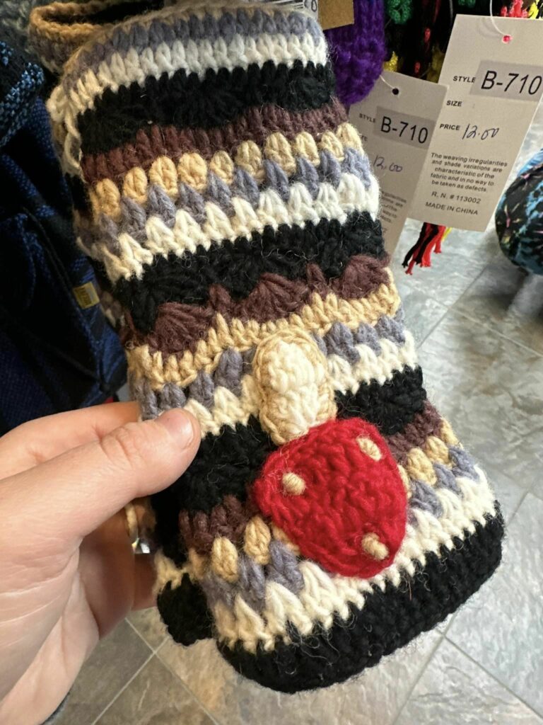 A knit sock with a knit red and white mushroom on the front. The sock is knit in colors of deep forest green, light gray, brown, dark tan and light tan.