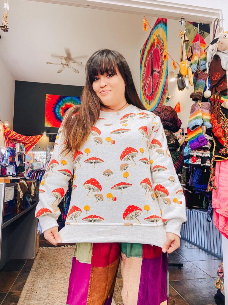 A woman shows off a white sweatshirt that she's wearing with red mushrooms, and other color mushrooms on it.  A variety of knit socks hang in the background.