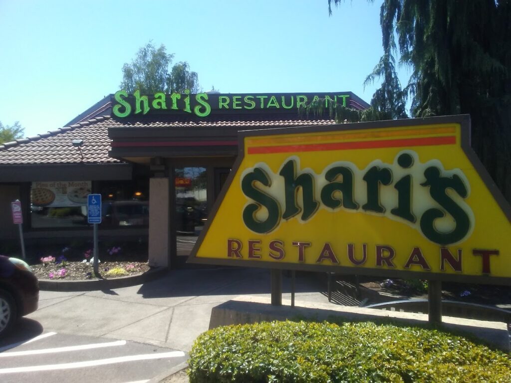 The outside of Sharis restaurant. This one has a yellow sign with green letters and accents of brown. There's a light up neon green sign on the building.