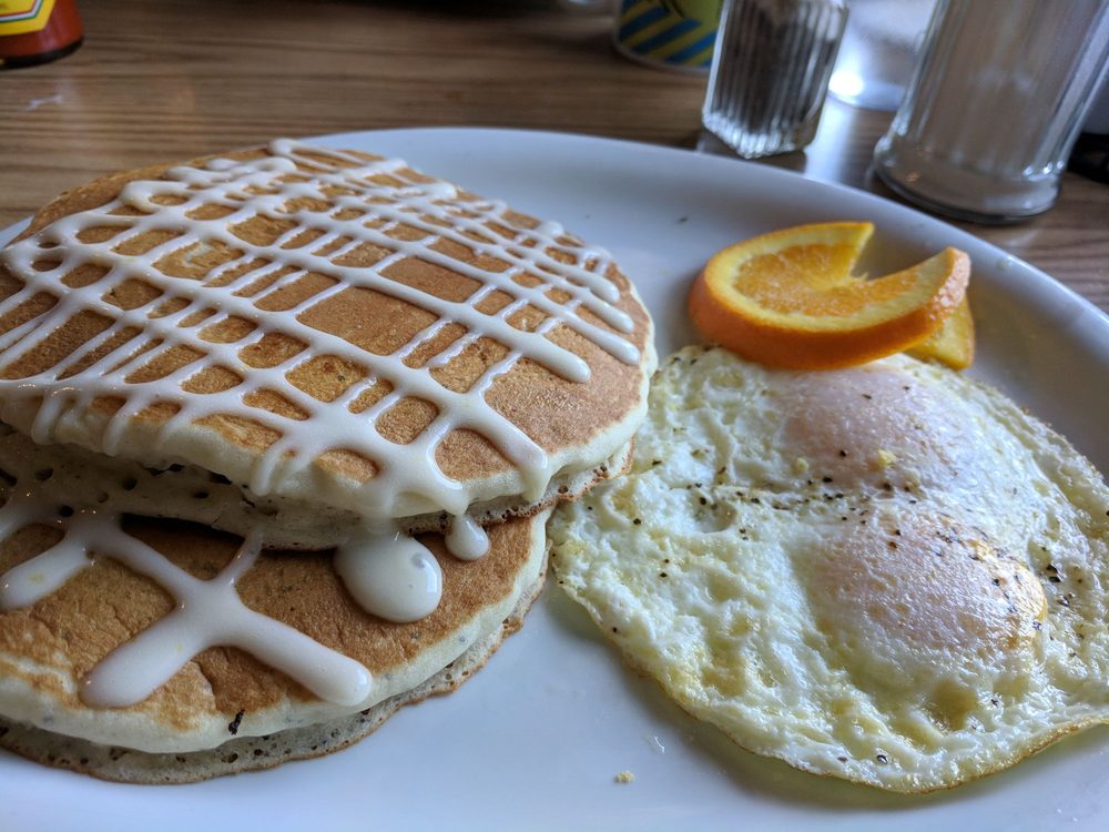 Pancakes drizzled in white sauce, next to two fried eggs and an orange slice. Looks good!