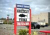 The sign for Grizzly Tuna. The sign is white with black words and a black silhouette of a tuna fish. It says "Locally Owned, Tuna Fish & Chips." The sign is held up by two bright red supports.