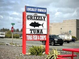 The sign for Grizzly Tuna. The sign is white with black words and a black silhouette of a tuna fish. It says "Locally Owned, Tuna Fish & Chips." The sign is held up by two bright red supports.