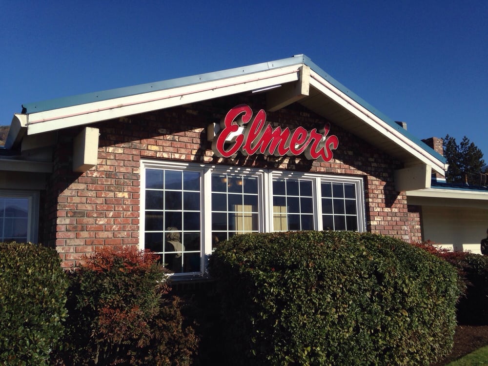The outside of an Elmer's Restaurant.  The restaurant is brick with bushes in front of a large window.  There's a red Elmer's sign above the windows on the side of the building.  The sign is in cursive.