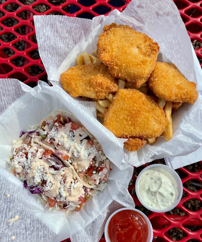 Fish and Chips , and two fish tacos in takeout containers sitting on a red metal picnic table. Two small containers of sauce sit next to the food.