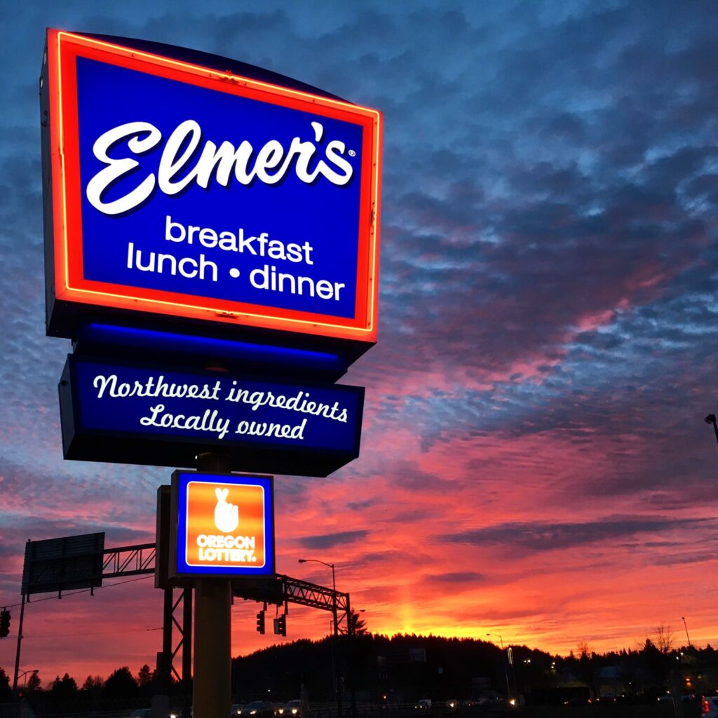 A blue and red Elmer's sign lit up as the sky grows dark with bright red, orange, pink and yellow colors from the sunset in the background.  There are interesting looking clouds in the sky behind the sign.  The sign reads: Elmer's - Breakfast, lunch, dinner.