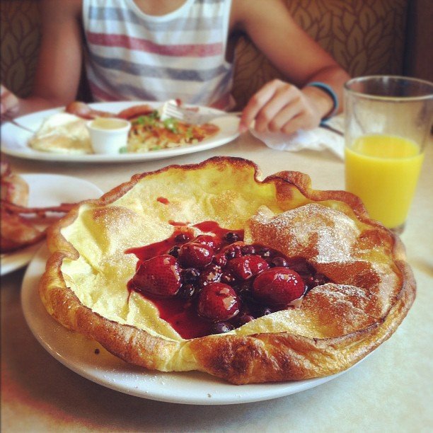 A huge German pancake with red berries on top. A glass of orange juice sits on the table next to it, and person eats eggs and hash browns in the background.