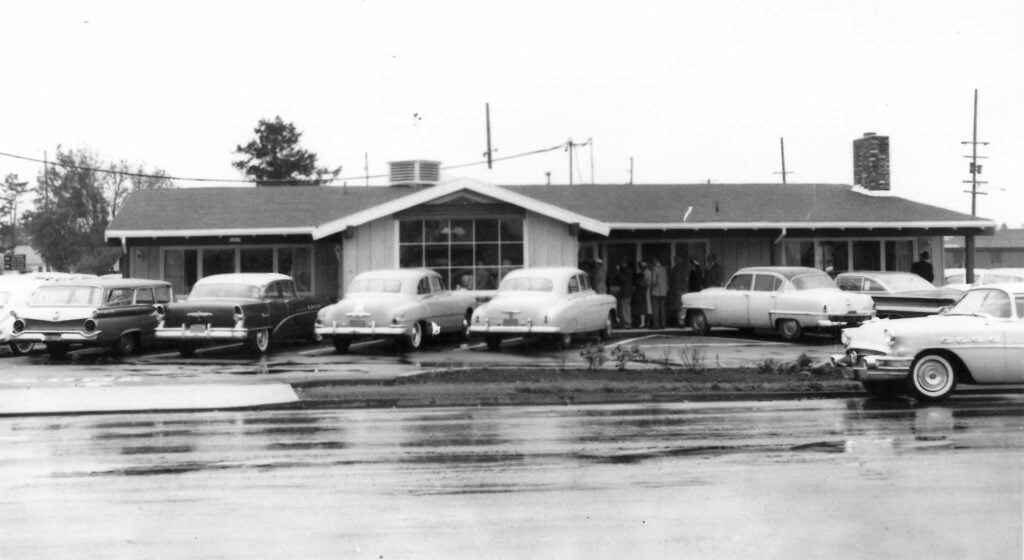 A black and white photo of Elmer's Restaurant in 1962. There are several cars parked outside and people heading inside.