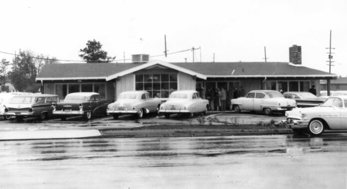 A black and white photo of Elmer's Restaurant in 1962. There are several cars parked outside and people heading inside.