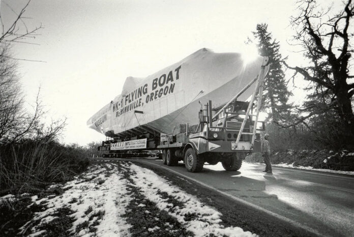 A black and white photo of the fuselage of the Spruce Goose being transported down the road towards McMinnville, Oregon.