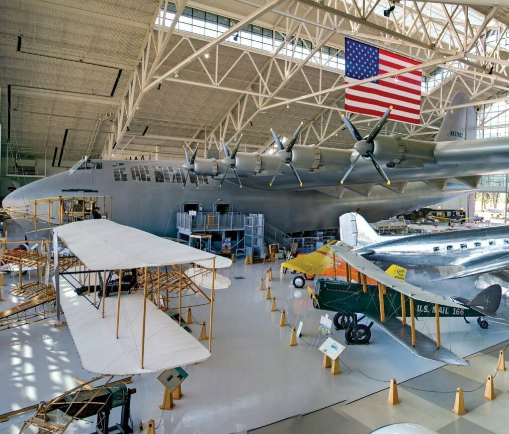 The massive Spruce Goose inside the Evergreen Aviation And Space Museum. It's painted silver or gray and there are other historical aircraft surrounding it.