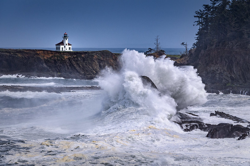The Cape Arago Lighthouse with a huge wave crashing against a rock in the foreground.
