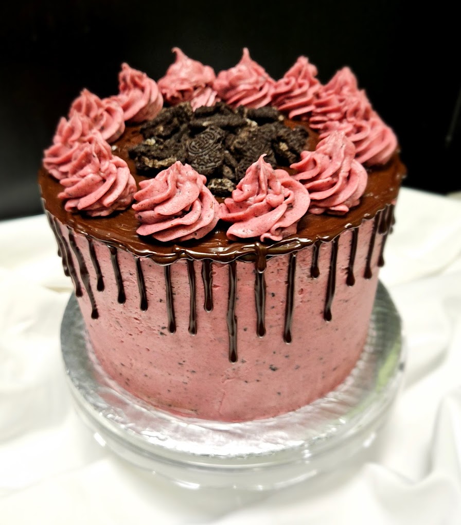 A crazy looking photo of a cake. It's a pink cake with chocolate drizzled on top and down the side. There's pink frosting piped on top and crumbled Oreo cookies in the center of the pink piped frosting.