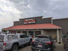 The outside of Roosters Country Kitchen in Pendleton, Oregon. It's a gray building with a red roof and red and white sign. It has farm vibes. There are two cars parked out front in the parking lot and a gray cloudy sky.