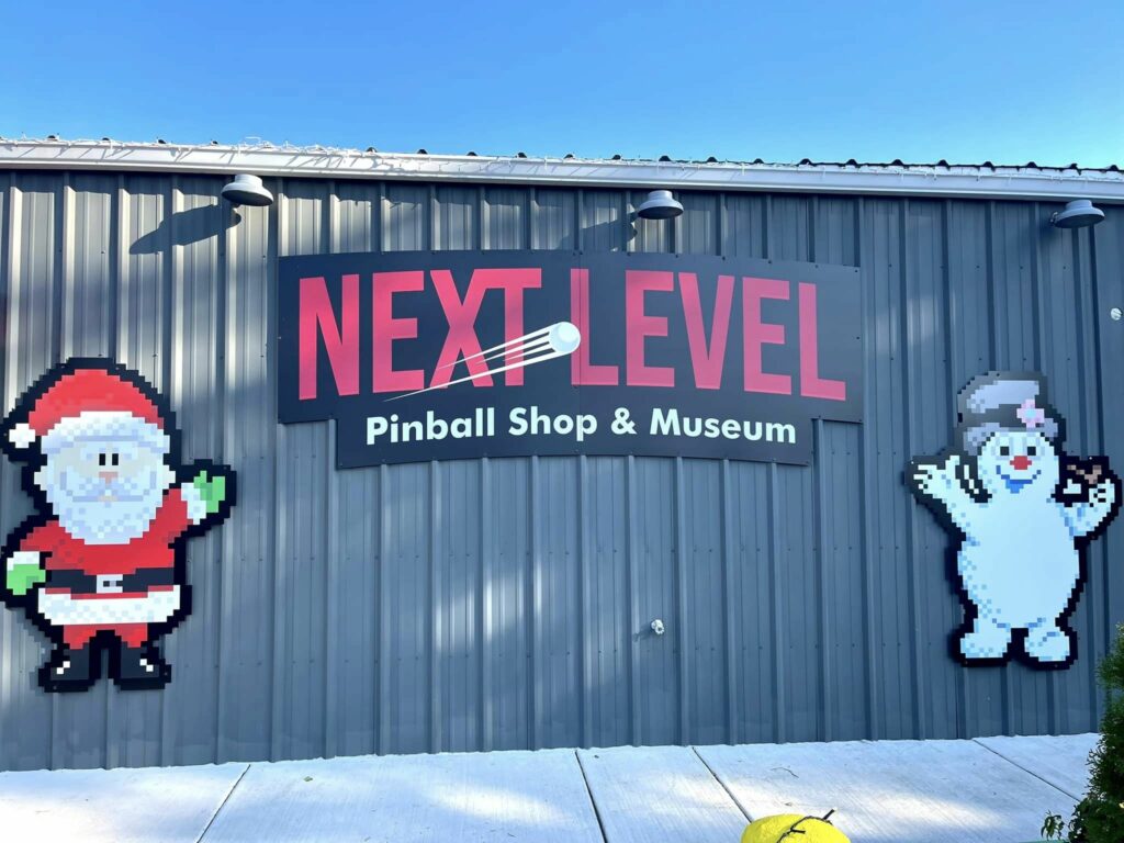 The outside of Next Level Pinball. The sign is bright red, and there's cartoon characters next to it. The building has gray metal siding.