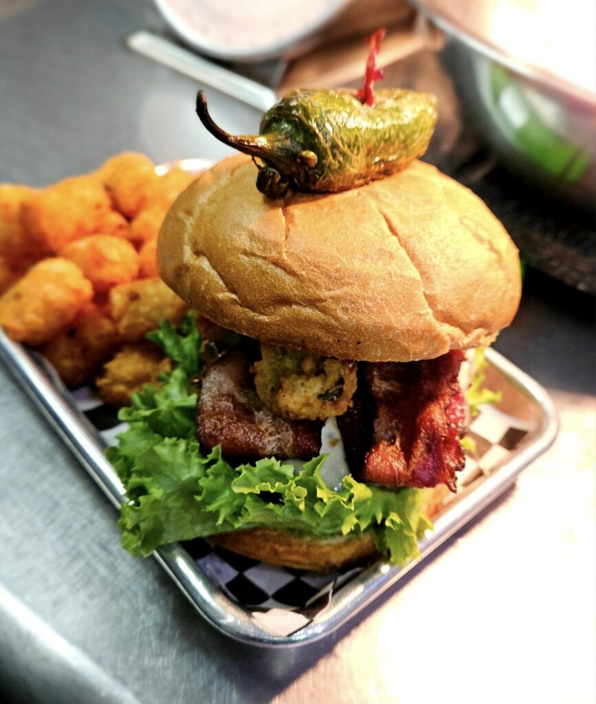 St Helen's Inferno Burger. It's got a toothpick sticking out the top and holding a fried jalapeno in place. This burger looks massive and loaded with toppings. There are tater tots in the background.