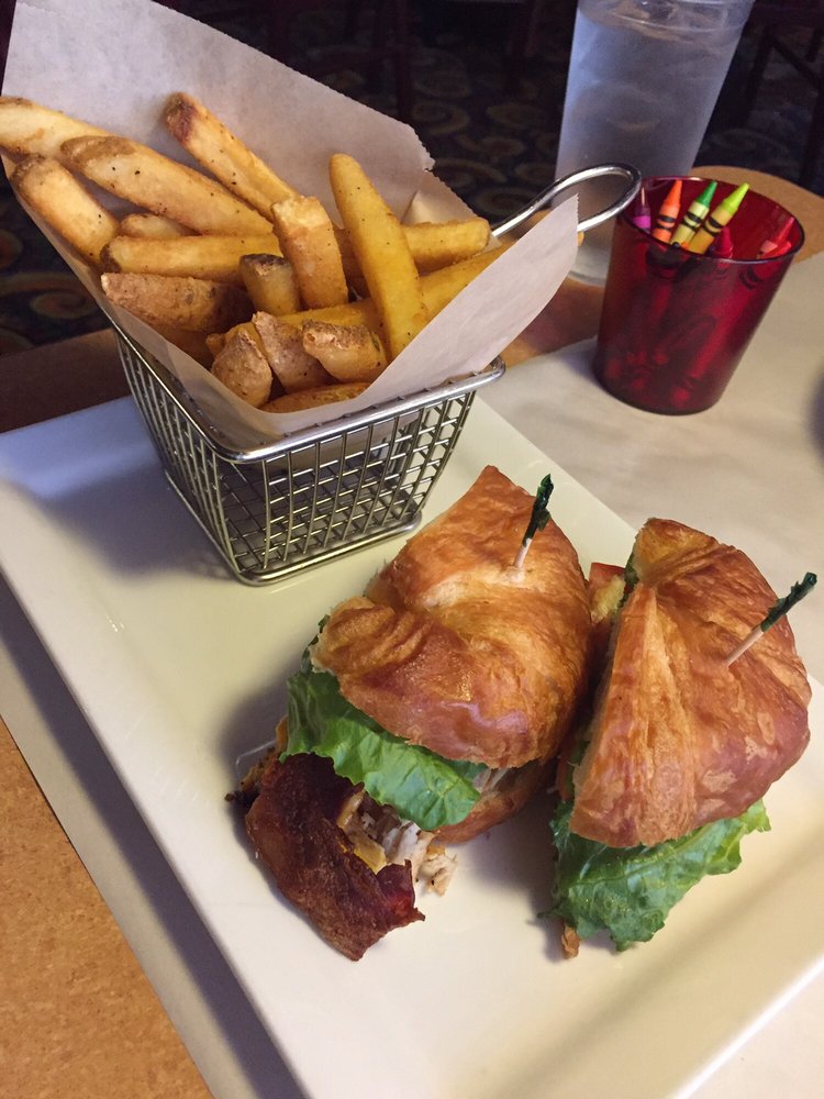 A turkey bacon club croissant sandwich with a metal basket of French fries. There is also a red plastic cup of crayons sitting on the table.